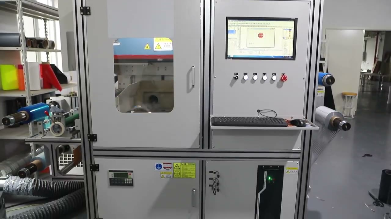 Still from factory video: an enclosed laser cutting machine with control panel and material rolls on the floor